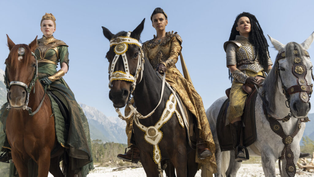 Three regal women in elaborate fantasy armor ride horses side by side against a clear blue sky and mountainous backdrop. The woman on the left wears a green gown and golden bracers, riding a chestnut horse adorned with silver. The center woman wears ornate gold robes with matching armor, mounted on a dark horse decorated with white and gold harnesses. On the right, a woman in silver and gold armor with long black locks rides a white horse decked with red and gold embellishments.