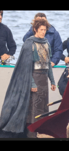 Leaked set photo showing an Elven character in a long cloak and a breastplate standing on a rocky promontory. Crew members are standing around. The actor playing the elf is not one of the leading actors.