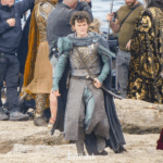 Robert Aramayo as Elrond in a leaked set photo walking towards the camera, wearing a long robe and a breastplate, as well as as sword at his side. Gil-galad and Ar-Pharazôn can be partially seen in the background, as well as crew members.