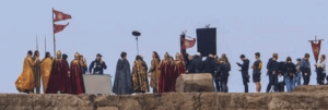 Wide shot of a group of actors in costumes and crew members on a rocky platform with flags and banners. There appears to be a mix of Númenórean and Elven guards, as well as what appears to be Elrong, Ar-Pharazôn and Gil-galad standing together in the middle