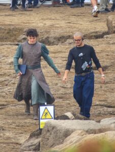 Robert Aramayo as Elrond holding an iPad and walking towards camera with a crew member, in a leaked set photo. The ground is rocky and there is a warning sign.