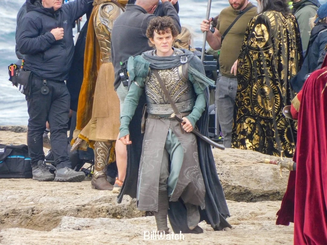 Robert Aramayo as Elrond in a leaked set photo walking towards the camera, wearing a long robe and a breastplate, as well as as sword at his side. Gil-galad and Ar-Pharazôn can be partially seen in the background, as well as crew members.