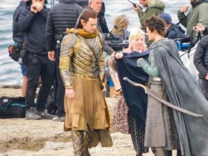 Ben Walker as Gil-galad looks on as a crew member holds a poncho for Rob Aramayo's Elrond, who is equipped with a sword. Crew members stand around in the background.