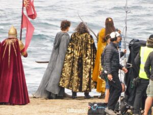 Elrond, Ar-Pharazôn and Gil-galad stand on a rocky platform looking out to sea, while accompanied by a guard in a red cloak holding a flag. Crew members are standing alongside. Gil-galad is holding his spear Aeglos.