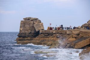 Extra wide shot of a rocky promontory surrounded by see with cast and crew standing in groups along its length