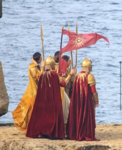 Guards in robes, carrying spears and a flag. The flag is red with a sun-logo. The guards with red robes also have a sun motif, while the guards with golden robes have an Elven pattern on theirs. These latter actors are wearing hair-nets.
