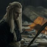 Charlie Vickers as Sauron from The Rings of Power, holding a spear and looking at its point, against the background of the ruined remains of Celebrimbor's forge.
