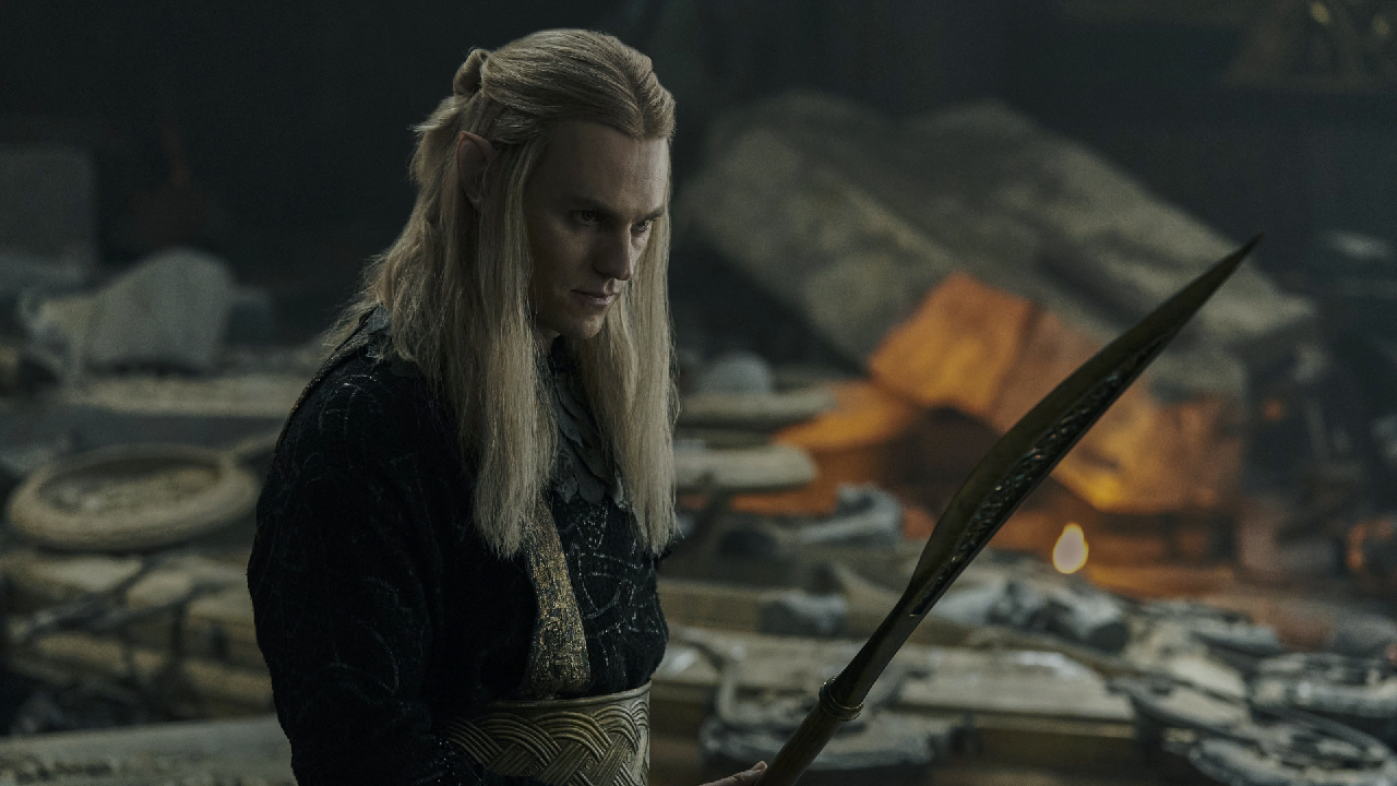 Charlie Vickers as Sauron from The Rings of Power, holding a spear and looking at its point, against the background of the ruined remains of Celebrimbor's forge.