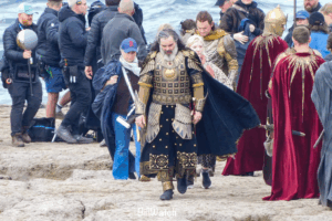 Trystan Gravelle walking towards the camera in an elaborate black and gold costume, with breastplate, and armour on this thighs, forearms, shins and shoulders. Crew members are also standing around. Ben Walker as Gil-galad can be seen behind him, as well as guards in long red cloaks with a golden sun motif. The ground is rocky and the sea is visible in the background
