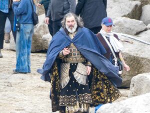 Trystan Gravelle as Ar-Pharazôn walking towards the camera over rocky ground in a leaked set photo. He is wearing a blue poncho over his elaborate black and gold costume. Crew members are standing around too.