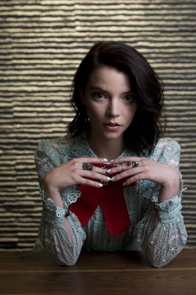 Portrait of Anya Taylor-Joy with very dark brown hair, leaning forward on a table with her elbows while her fingers are linked in front of her.