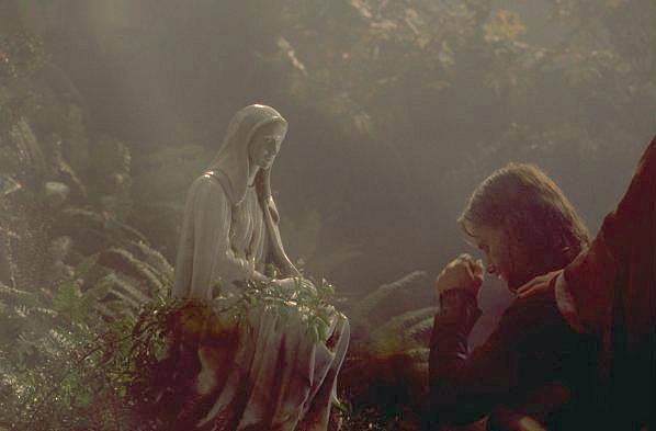 Moody shot of a statue of a hooded woman (Gilraen) in a forest. A man (Aragorn) is kneeling in front of it respectfully, his head bowed, while a hand of a person out of shot is resting on his shoulder