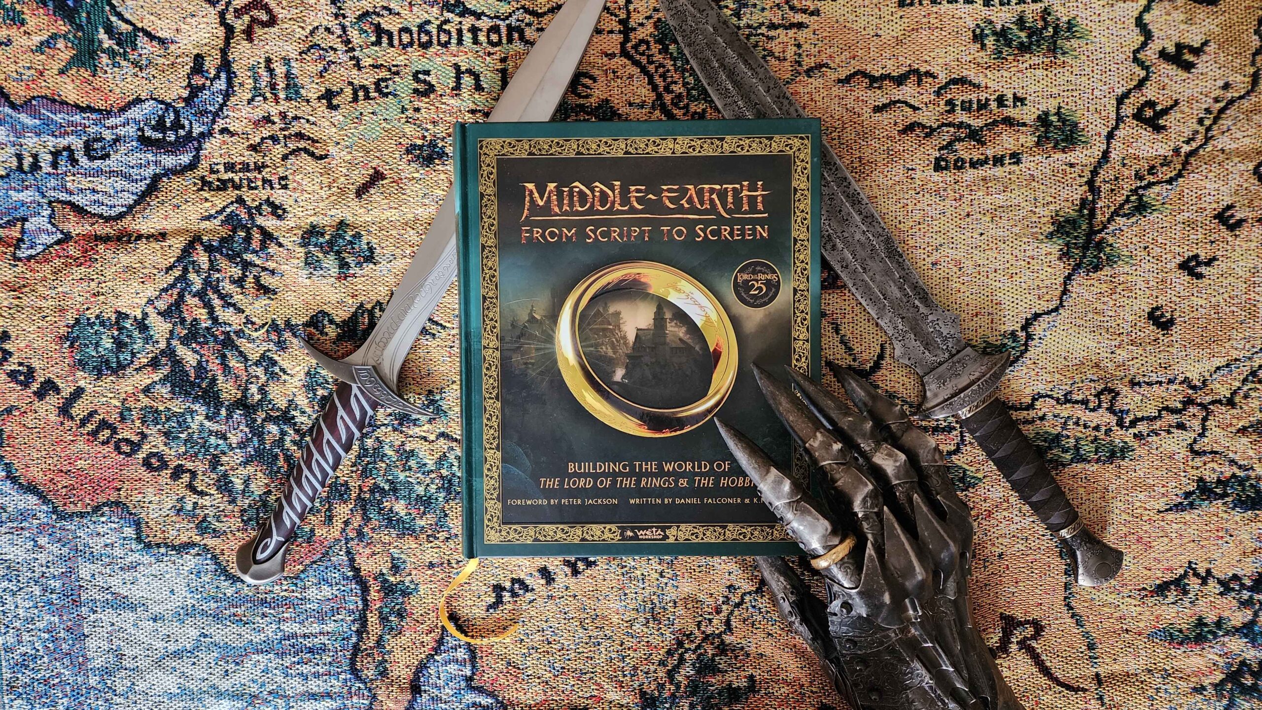 A photo of the front cover of Middle-earth from Script to Screen, on a cloth map of Middle-earth. There are replica movie props either side: two swords and Sauron's gauntlet.