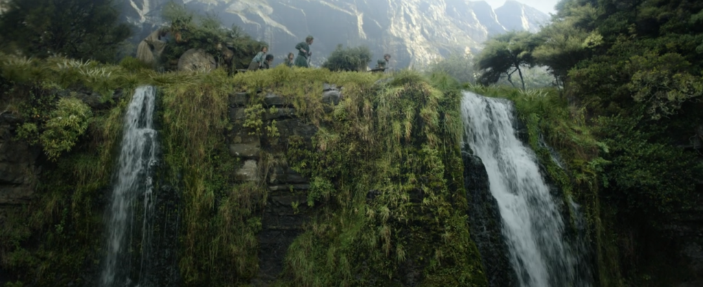 Landscape shot from low down in front of a waterfall looking up at the Brandyfoot family and the Stranger pushing a caravan along the top of the cliff