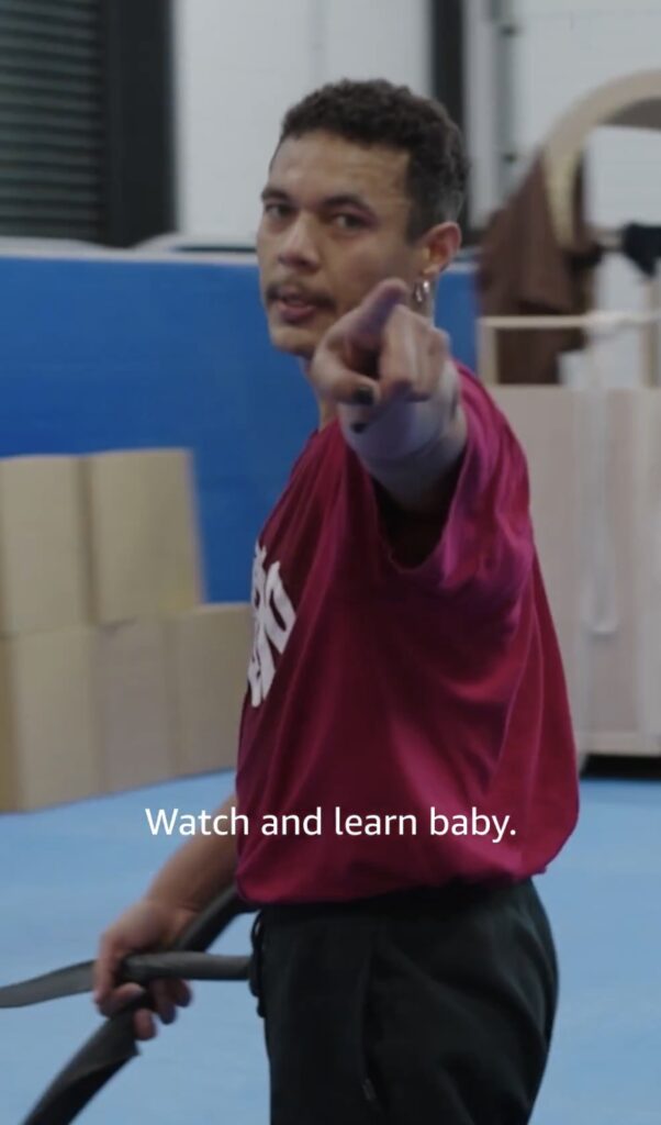 Ismael Cruz-Córdova in a practice gym holding a prop weapon, looking and pointing direct to camera with subtitle "Watch and learn baby"