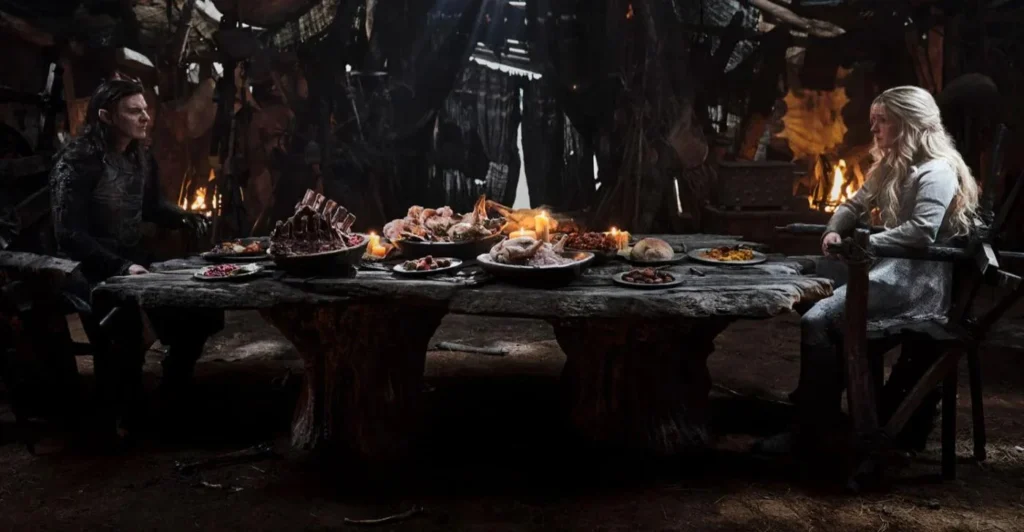 Adar and Galadriel sitting at opposite ends of a long rough table laden with food in a makeshift tent. Galadriel looks very uncomfortable.