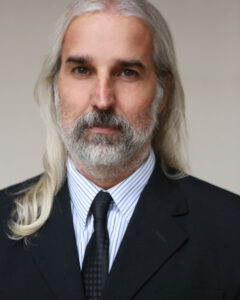 Promotional head and shoulders photo of Pablo Sciepura wearing a suit and tie. He has long grey hair, and a trimmed beard that is a mix of black and white