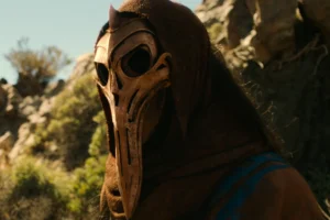 Kilta the Rhûnic tracker in Rings of Power in an outdoors scene. He is wearing a metal mask and hood.