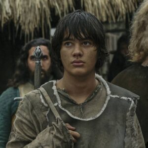 Tyroe Muhafidin as Theo in Rings of Power. He is carrying a sword slung over his shoulder by a strap and surrounded by some other Southlanders in Pelargir