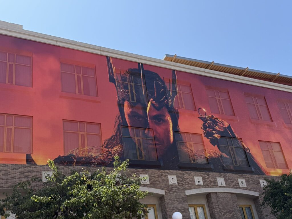 Photo of a wrapped building with a giant front-on photo of Sauron's head and shoulders, with long dark hair. He is wearing a the re-tooled Morgoth crown, and holding up a gold ring in a gauntleted hand. The background is red and smoky
