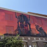 Photo of a wrapped building with a giant front-on photo of Sauron's head and shoulders, with long dark hair. He is wearing a the re-tooled Morgoth crown, and holding up a gold ring in a gauntleted hand. The background is red and smoky