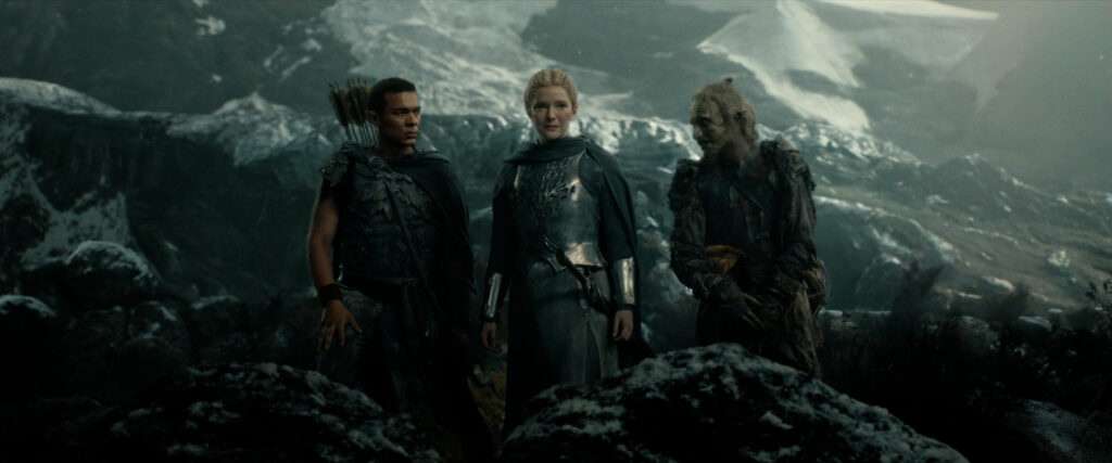 The elves Arondir and Galadriel with the Orc Marnûkh standing on a rocky outcrop