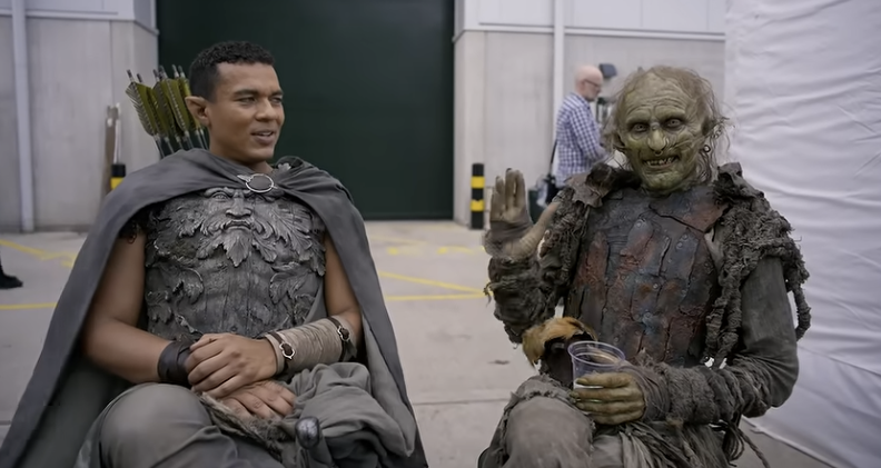 Ismael Cruz Córdova and Adam Young sitting on seats in costume on a filming back lot. Adam is holding a plastic cup of water, smiling broadly and waving to the camera, rather incongruous for his orc prosthetics