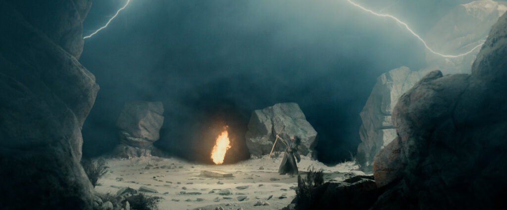Gandalf with his staff raised against a strong wind, in the middle of a rocky circle with a magical flame emerging from a rock, and lightning around him